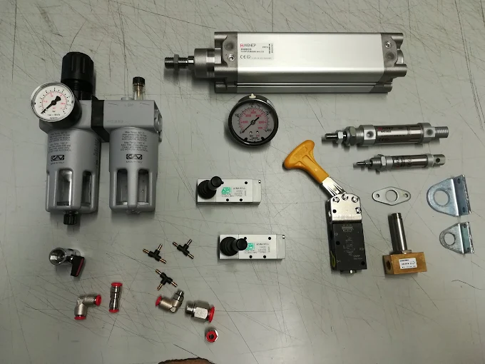 Componenti pneumatici su piano grigio: filtro regolatore con manometro, cilindri in alluminio, elettrovalvole e raccordi rossi